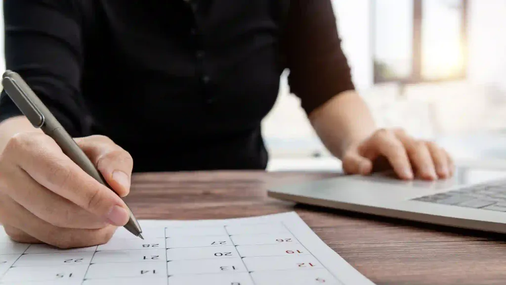 Persona marca fechas en calendario impreso sobre escritorio con laptop y luz natural