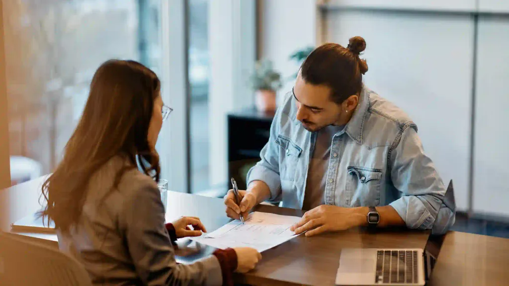 Joven emprendedor firmando documento sobre escritorio acompañado por asesora profesional