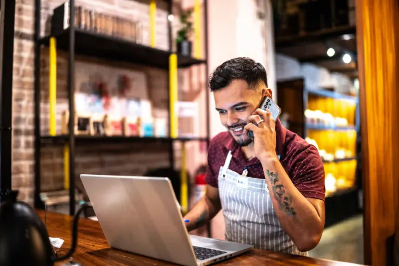 Emprendedor con delantal atiende llamada telefónica junto a laptop en mostrador de tienda pequeña