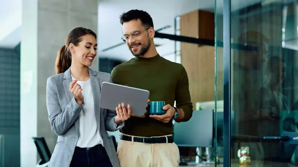 Pareja de profesionales sonríe mientras revisa información en tablet en oficina moderna con paredes de vidrio
