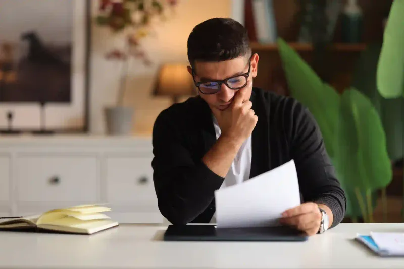 Empresario joven con anteojos revisa documento tributario en gesto pensativo sobre escritorio