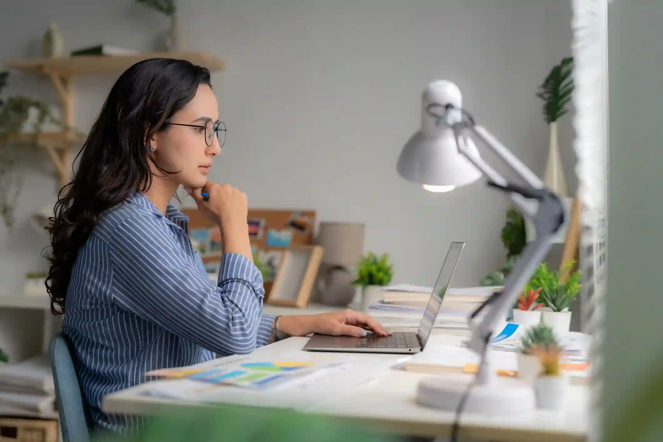 Mujer profesional con anteojos analiza información en laptop sobre escritorio con plantas y lámpara