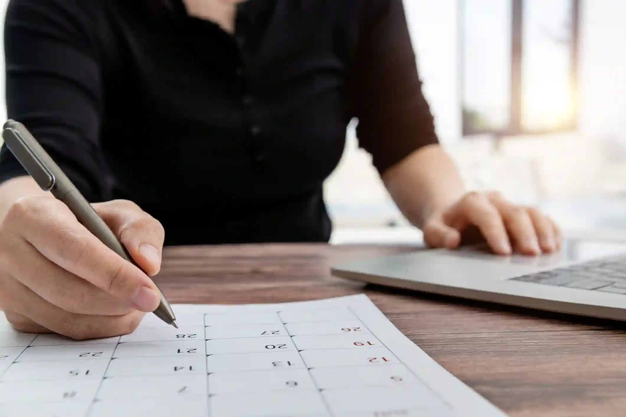 Persona marca fechas en calendario impreso sobre escritorio con laptop y luz natural